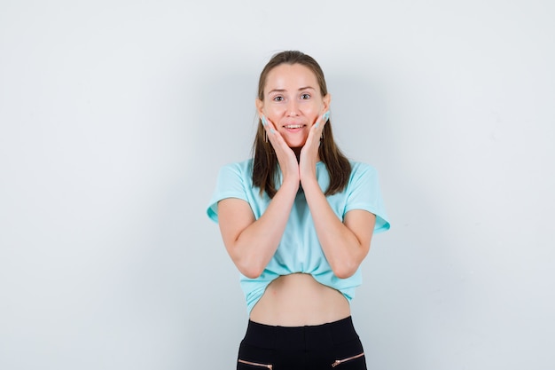 Portrait of young beautiful female with hands on cheeks in t-shirt and looking merry front view