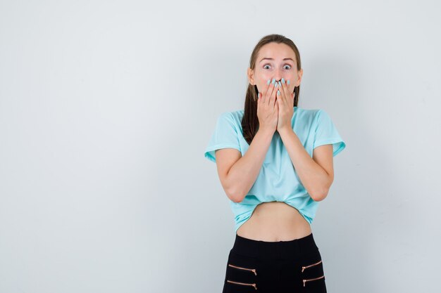 Portrait of young beautiful female with hands on mouth in t-shirt, pants and looking shocked front view