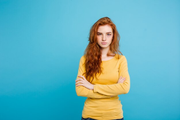 Portrait of young beautiful ginger woman with tender serious face crossing arms looking at camera. Isolated on pastel blue background. Copy space.