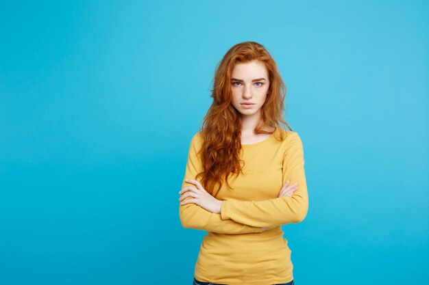 Portrait of young beautiful ginger woman with tender serious face crossing arms looking at camera. Isolated on pastel blue background. Copy space.