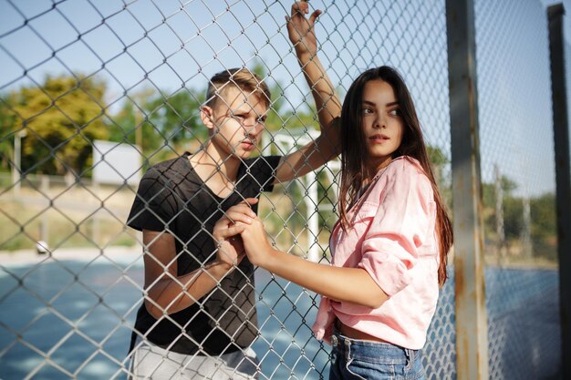 Portrait of young beautiful girl and boy standing between mesh fence on basketball court and thoughtfully looking aside