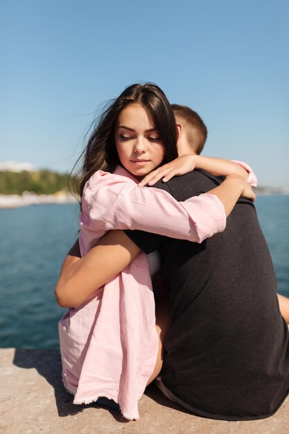 Portrait of young beautiful girl sitting and embracing cool boy while thoughtfully looking aside with sea view on background