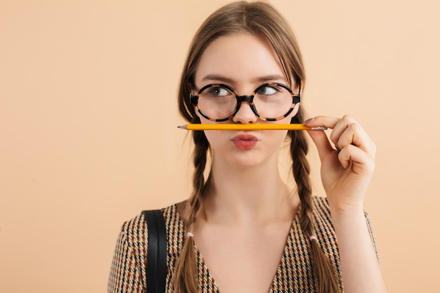Портрет молодой красивой девушки с двумя косами в современных очках, держащей карандаш над губами и мечтательно смотрящей в сторону на бежевом фоне