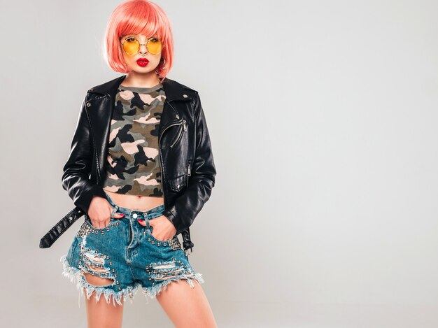 Portrait of young beautiful hipster bad girl in black leather jacket and earring in her nose.Sexy carefree smiling woman sitting in studio in pink wig near blue wall.Confident model in sunglasses