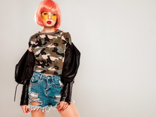 Free photo portrait of young beautiful hipster bad girl in black leather jacket and earring in her nose.sexy carefree smiling woman sitting in studio in pink wig near blue wall.confident model in sunglasses