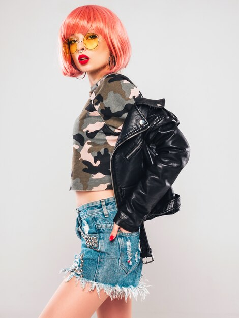 Portrait of young beautiful hipster bad girl in black leather jacket and earring in her nose.Sexy carefree smiling woman sitting in studio in pink wig near gray wall.Confident model in sunglasses