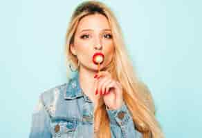 Free photo portrait of young beautiful hipster bad girl in trendy jeans  clothes and earring in her nose.  positive model licking round sugar candy