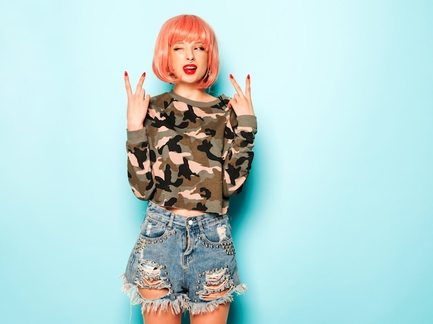 Portrait of young beautiful hipster bad girl in trendy jeans shorts and earring in her nose.Sexy carefree smiling woman posing in studio in pink wig.Positive model having fun.Shows peace sign