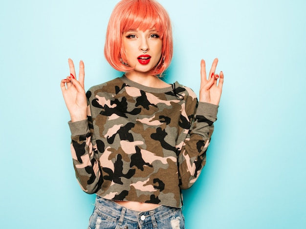 Portrait of young beautiful hipster bad girl in trendy jeans shorts and earring in her nose.sexy carefree smiling woman posing in studio in pink wig.positive model having fun.shows peace sign