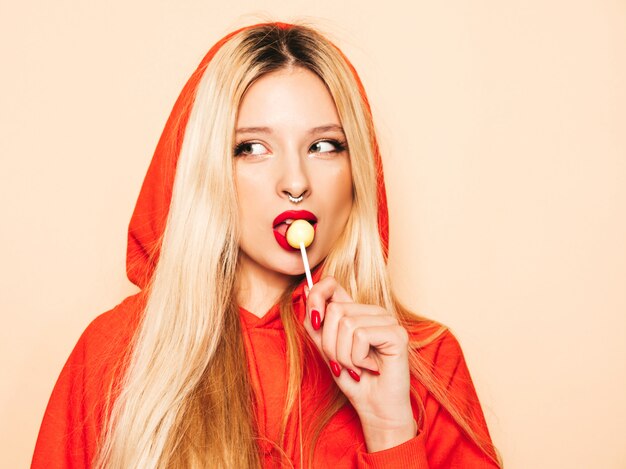 Portrait of young beautiful hipster bad girl in trendy red  hoodie and earring in her nose.  Positive model licking round sugar candy