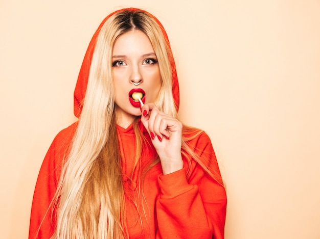 Portrait of young beautiful hipster bad girl in trendy red  hoodie and earring in her nose.  Positive model licking round sugar candy