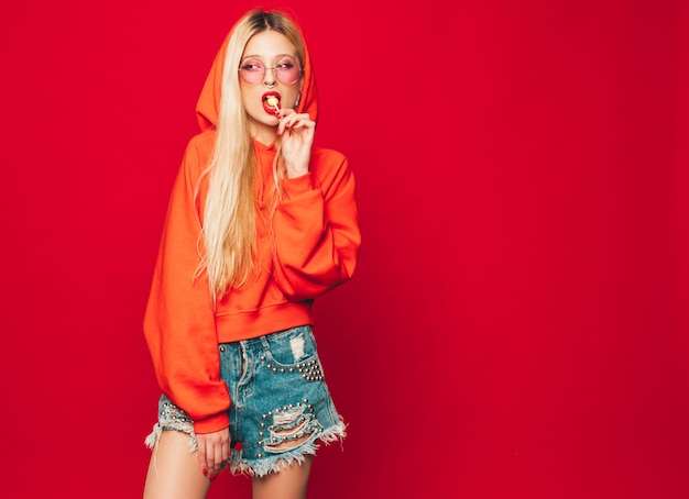 Portrait of young beautiful hipster bad girl in trendy red  hoodie and earring in her nose.  Positive model licking round sugar candy