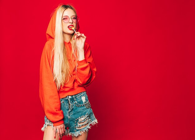 Portrait of young beautiful hipster bad girl in trendy red  hoodie and earring in her nose.  Positive model licking round sugar candy