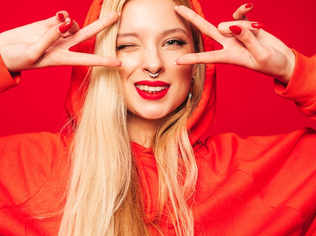 Portrait of young beautiful hipster bad girl in trendy red  hoodie and earring in her nose.  Positive model shows peace sign