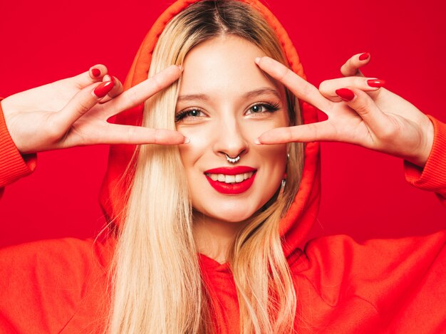 Portrait of young beautiful hipster bad girl in trendy red  hoodie and earring in her nose.  Positive model shows peace sign