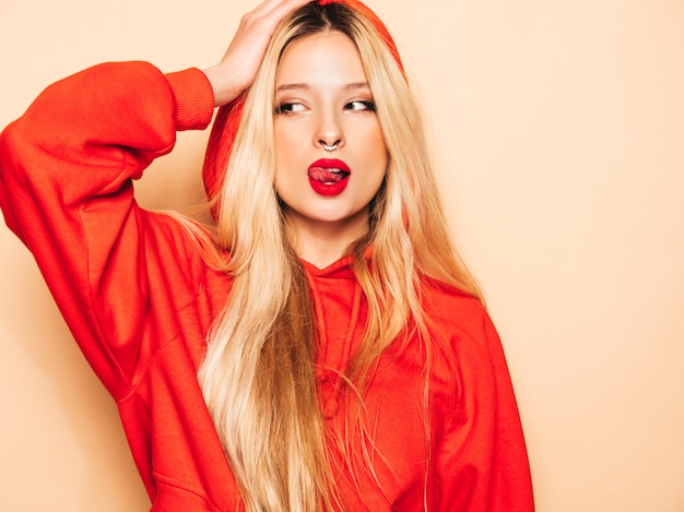 Portrait of young beautiful hipster bad girl in trendy red  hoodie and earring in her nose.  positive model shows tongue