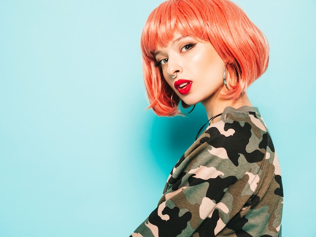 Free photo portrait of young beautiful hipster bad girl in trendy red summer clothes and earring in her nose.sexy carefree smiling woman posing in studio in pink wig near blue wall.positive model having fun