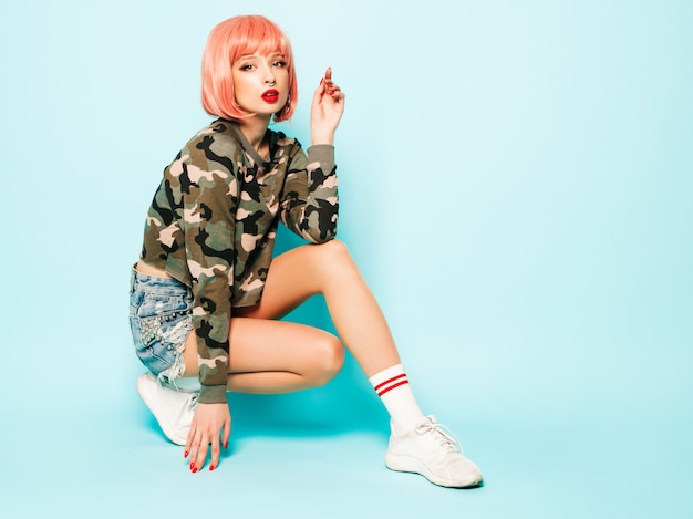 Portrait of young beautiful hipster bad girl in trendy red summer clothes and earring in her nose.Sexy carefree smiling woman sitting in studio in pink wig near blue wall.Positive model having fun