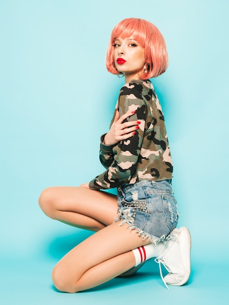 Free photo portrait of young beautiful hipster bad girl in trendy red summer clothes and earring in her nose.sexy carefree smiling woman sitting in studio in pink wig near blue wall.positive model having fun