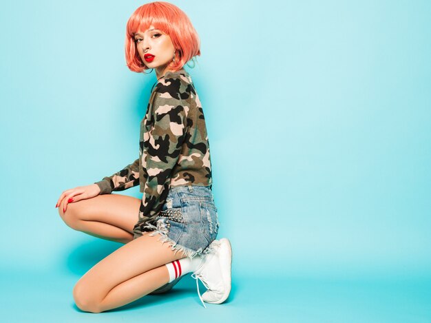 Portrait of young beautiful hipster bad girl in trendy red summer clothes and earring in her nose.Sexy carefree smiling woman sitting in studio in pink wig near blue wall.Positive model having fun