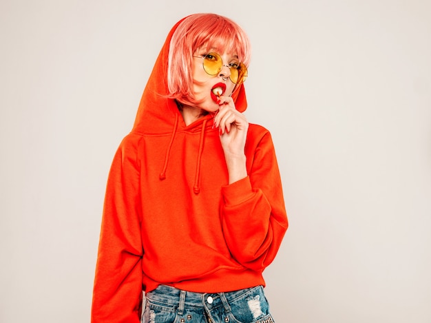 Portrait of young beautiful hipster bad girl in trendy red summer hoodie and earring in her nose.Sexy carefree smiling blond woman posing in studio in wig.Positive model licking round sugar candy