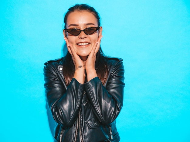 Portrait of young beautiful hipster  bad girl in trendy summer black leather jacket and sunglasses. Sexy carefree woman isolated on blue. Brunette model posing