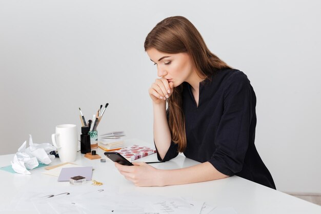 Portrait of young beautiful lady sitting at the white desk and thoughtfully looking in her cellphone isolated