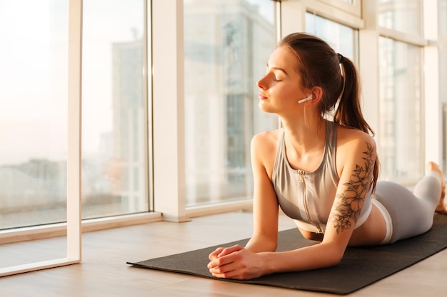 Portrait of young beautiful lady in sporty top and leggings lying on yoga mat and listening music in earphones while dreamily closing her eyes