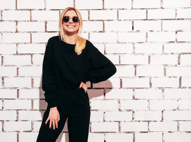 Portrait of young beautiful smiling blond female in trendy summer black oversize clothes Sexy carefree woman posing near white brick wall in studio Positive model having fun indoors In sunglasses