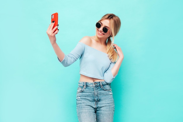 Portrait of young beautiful smiling blond female in trendy summer clothes Sexy carefree woman posing near blue wall in studio Positive model having fun indoors Cheerful and happy Taking selfie