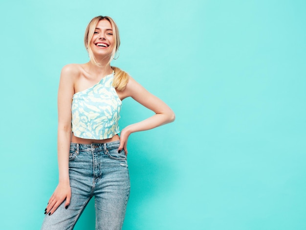 Портрет молодой красивой улыбающейся блондинки в модной летней одежде Сексуальная беззаботная женщина позирует возле синей стены в студии Позитивная модель веселится в помещении Веселая и счастливая