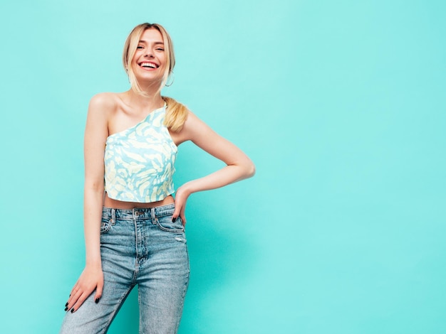 Portrait of young beautiful smiling blond female in trendy summer clothes Sexy carefree woman posing near blue wall in studio Positive model having fun indoors Cheerful and happy