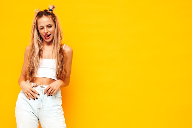 Portrait of young beautiful smiling blond female in trendy summer clothes Sexy carefree woman posing near yellow wall in studio Positive model having fun indoors Cheerful and happy Shows tongue