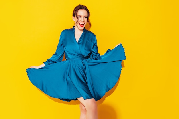 Portrait of young beautiful smiling female in trendy summer blue dress Sexy carefree woman posing near yellow wall in studio Positive model having fun and going crazy Cheerful and happy