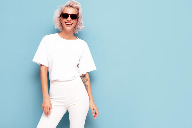 Portrait of young beautiful smiling female in trendy summer clothes Sexy carefree woman posing near blue wall in studio Positive blond model having fun and going crazy Cheerful and happy