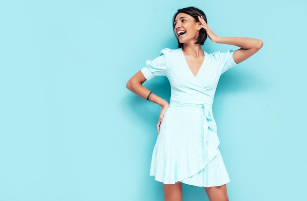 Портрет молодой красивой улыбающейся женщины в модном летнем платье Беззаботная женщина позирует у синей стены в студии Позитивная модель веселится в помещении Веселая и счастливая Изолированная
