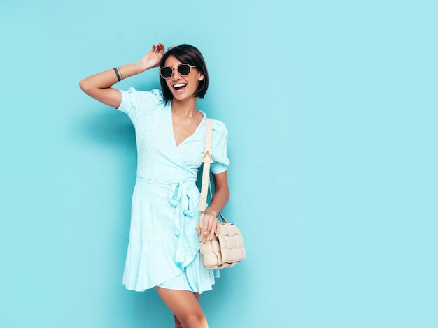 Portrait of young beautiful smiling female in trendy summer dress Carefree woman posing near blue wall in studio Positive model having fun indoors Cheerful and happy Isolated