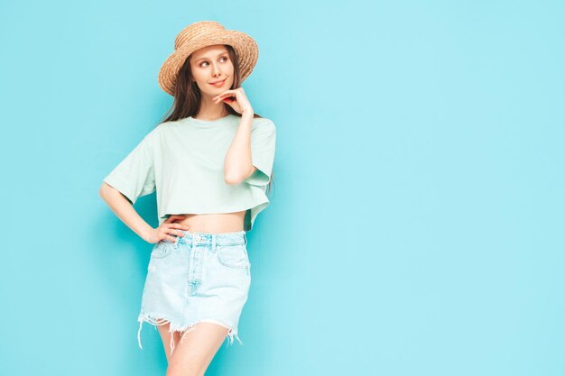 스튜디오의 파란색 벽 근처에서 포즈를 취한 트렌디한 여름 청바지 치마를 입은 평온한 여성의 초상화 실내에서 즐거운 시간을 보내는 긍정적인 모델 쾌활하고 행복한 모자