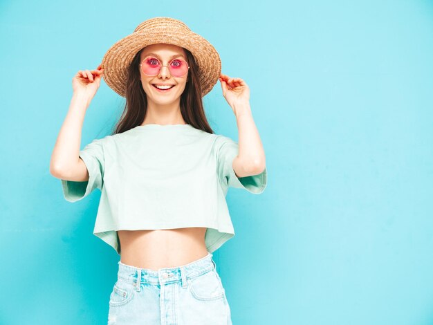 Портрет молодой красивой улыбающейся женщины в модной летней джинсовой юбке беззаботная женщина позирует возле синей стены в студии Позитивная модель веселится в помещении Веселая и счастливая В шляпе