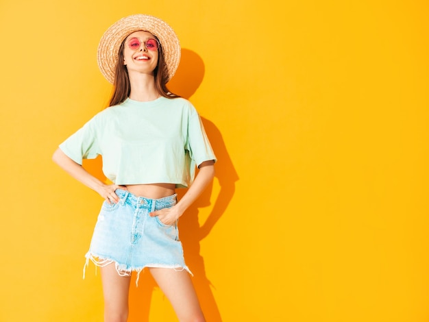 Portrait of young beautiful smiling female in trendy summer jeans skirt carefree woman posing near yellow wall in studio Positive model having fun indoors Cheerful and happy In hat