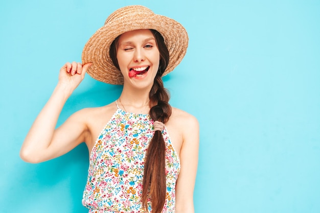 Portrait of young beautiful smiling female in trendy summer overalls carefree woman posing near blue wall in studio Positive model having fun indoors Cheerful and happy In hat