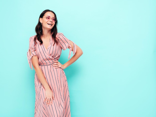 Portrait of young beautiful smiling female in trendy summer pink dress Sexy carefree woman posing near blue wall in studio Positive model having fun indoors Cheerful and happy in sunglasses