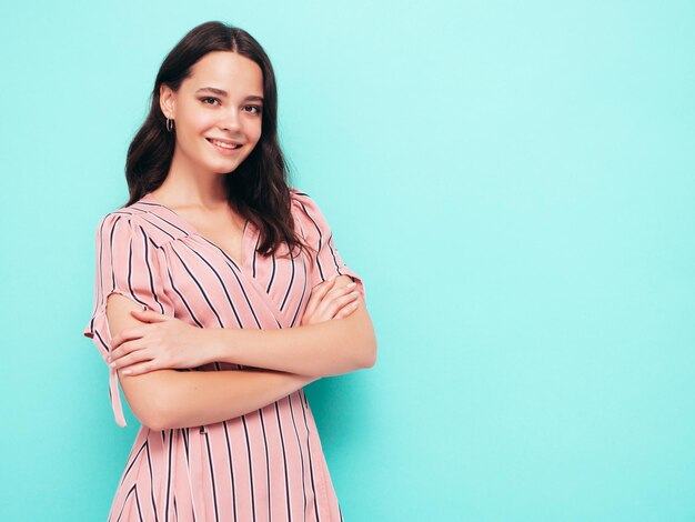 Portrait of young beautiful smiling female in trendy summer pink dress Sexy carefree woman posing near blue wall in studio Positive model having fun indoors Cheerful and happy