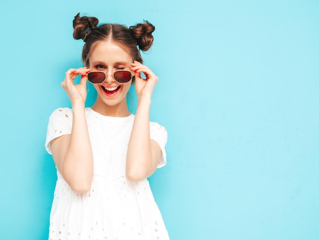 Foto gratuita ritratto di giovane bella donna sorridente in abito bianco estivo alla moda donna spensierata in posa vicino al muro blu in studio modello positivo che si diverte al chiuso allegro e felice si toglie gli occhiali da sole