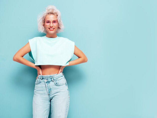 Portrait of young beautiful smiling female in trendy summer white dress Sexy carefree blond woman posing near blue wall in studio Positive model having fun indoors Cheerful and happy in sunglasses