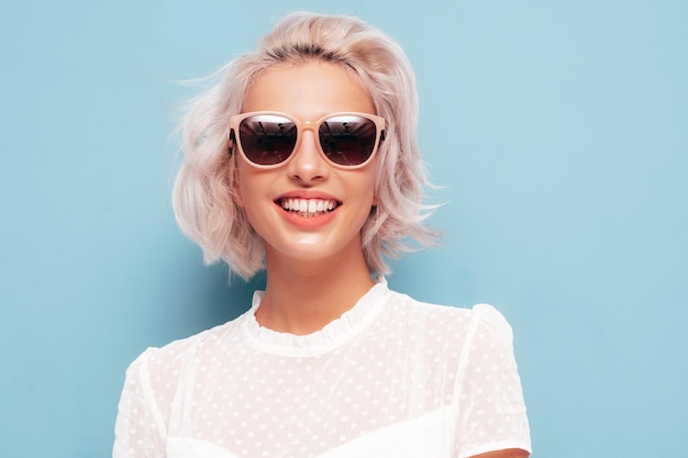 Портрет молодой красивой улыбающейся женщины в модном летнем белом платье Сексуальная беззаботная блондинка позирует у синей стены в студии Позитивная модель веселится в помещении Веселая и счастливая в солнечных очках