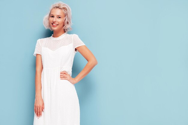 최신 유행의 여름 흰 드레스를 입은 젊은 아름다운 웃는 여성의 초상화 스튜디오의 파란색 벽 근처에서 포즈를 취하는 섹시하고 평온한 금발 여성 긍정적인 모델 실내에서 재미 명랑하고 행복합니다.