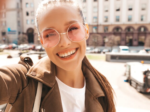 Portrait of young beautiful smiling girl in summer hipster jacket and jeans.Model taking selfie on smartphone.