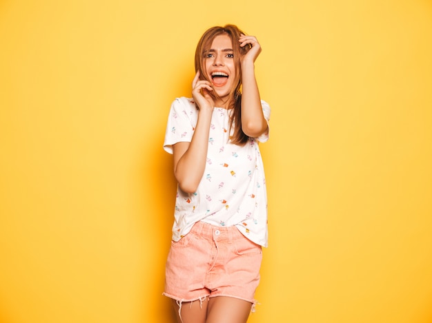 Портрет молодой красивой улыбающейся хипстерской девочки в модных летних джинсах шорты одевается. Сексуальная беззаботная женщина позирует возле желтой стены. Позитивная модель с удовольствием