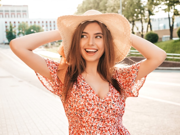 Портрет молодой красивой улыбающейся хипстерской девочки в модном летнем сарафане. Сексуальная беззаботная женщина, позирующая на уличном фоне в шляпе на закате. Позитивная модель на открытом воздухе